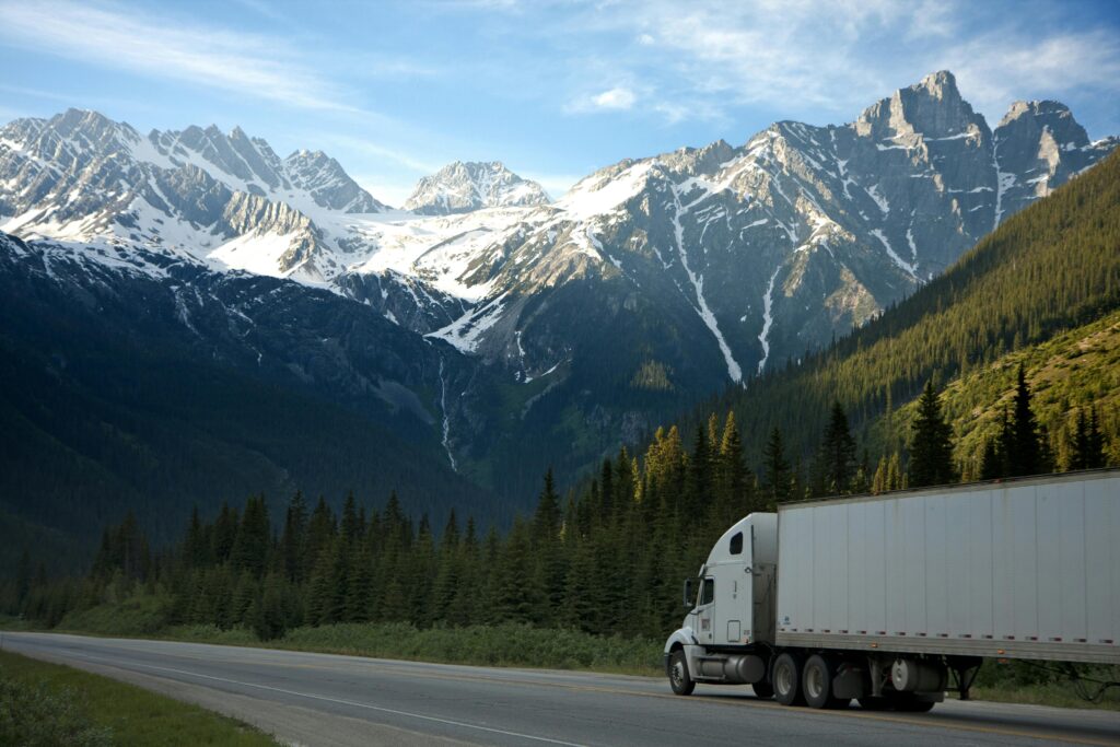 A semi-truck travels along a highway with snow-capped mountains in the background.